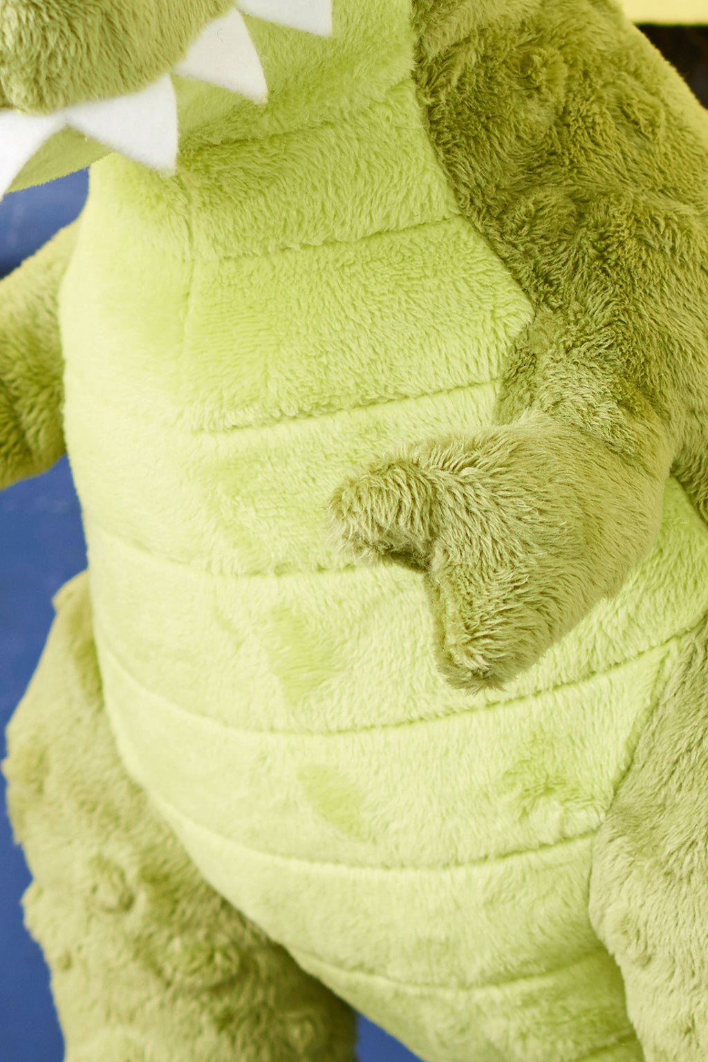 Close-up of a green plush dinosaur toy with a soft texture. It has white felt teeth and visible stitching lines, creating a playful and cuddly appearance.