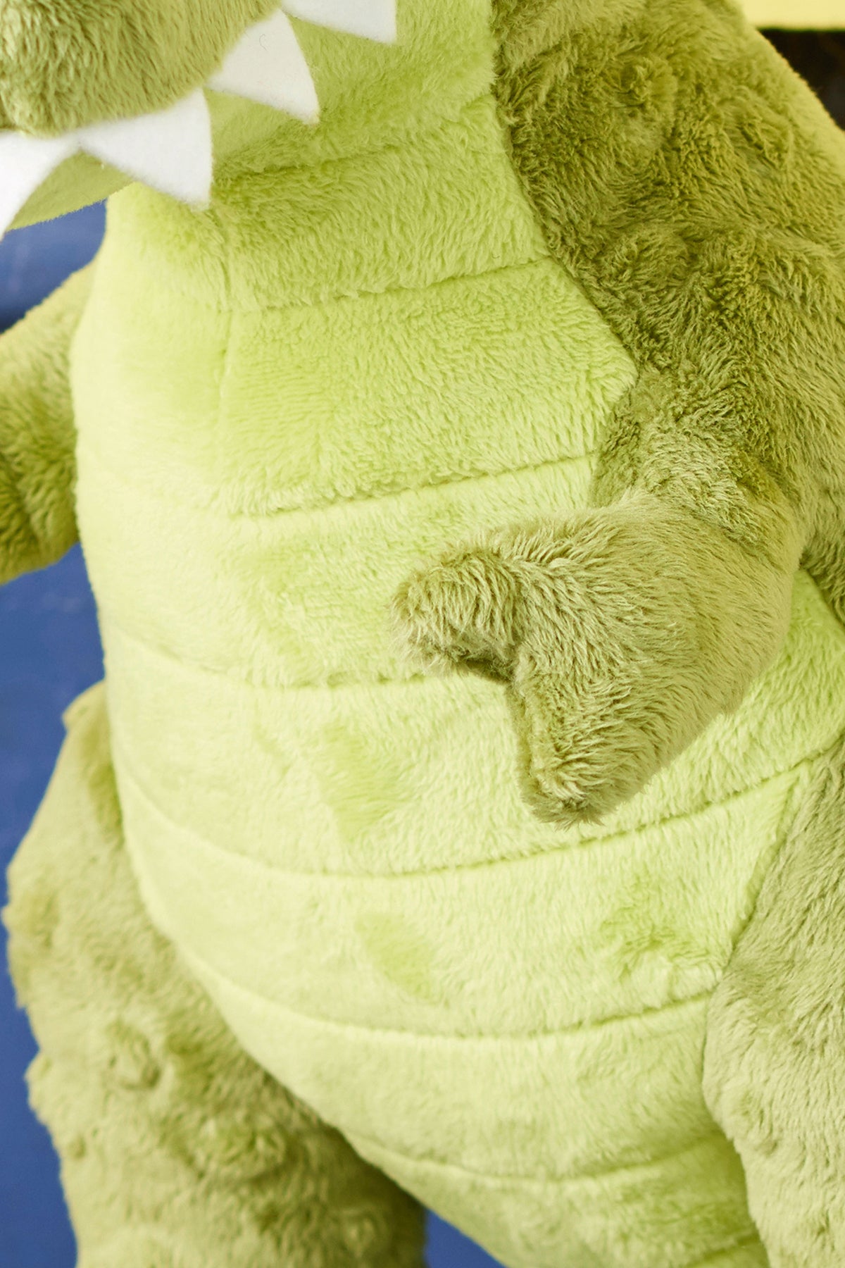 Close-up of a green plush dinosaur toy with a soft texture. It has white felt teeth and visible stitching lines, creating a playful and cuddly appearance.