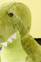 Close-up of a green plush dinosaur against a yellow background. The toy shows a textured surface, white felt teeth, and a glossy black eye, conveying a playful tone.