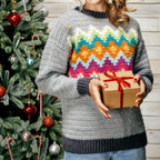 Person wearing a colorful sweater holding a gift box in front of a decorated Christmas tree.