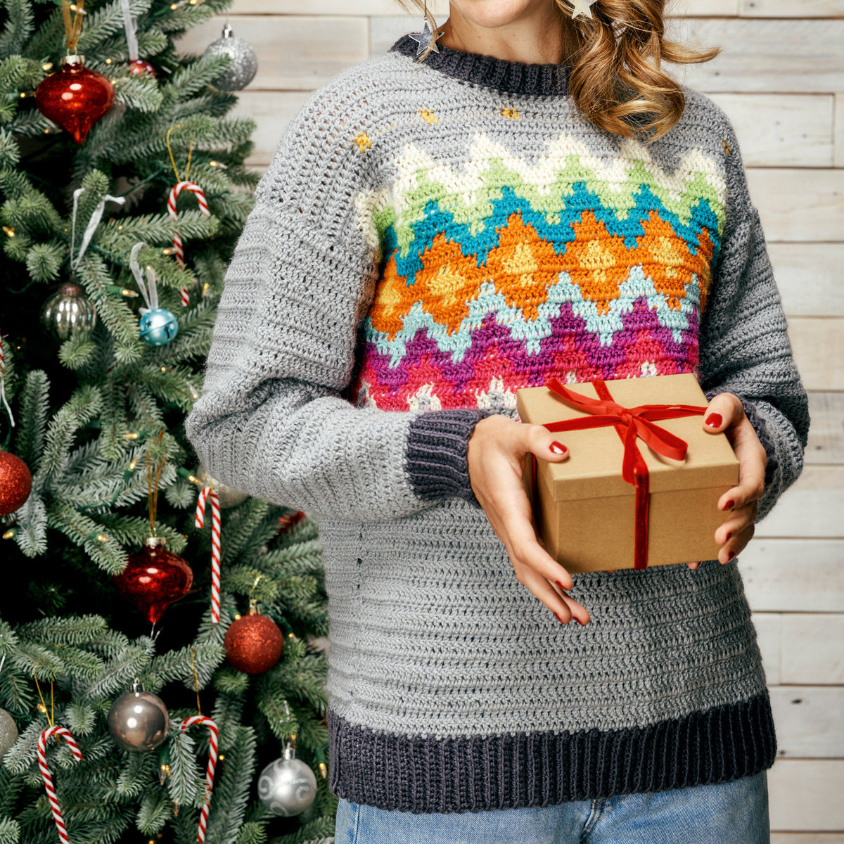 Person wearing a colorful sweater holding a gift box in front of a decorated Christmas tree.