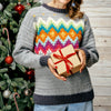 Person wearing a colorful patterned sweater holding a gift box in front of a Christmas tree.