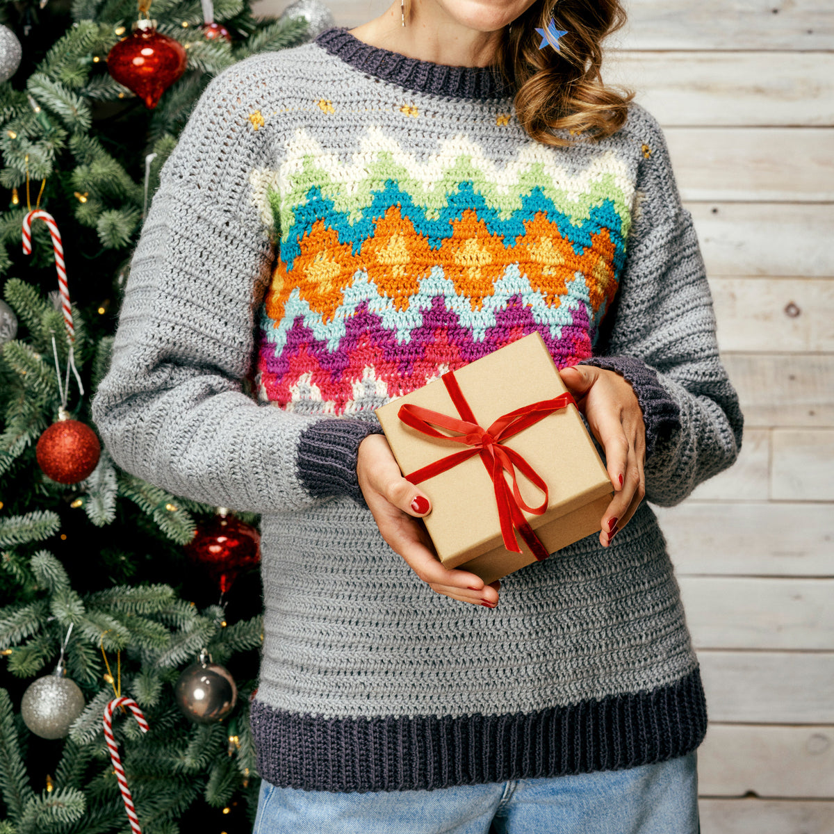Person wearing a colorful patterned sweater holding a gift box in front of a Christmas tree.