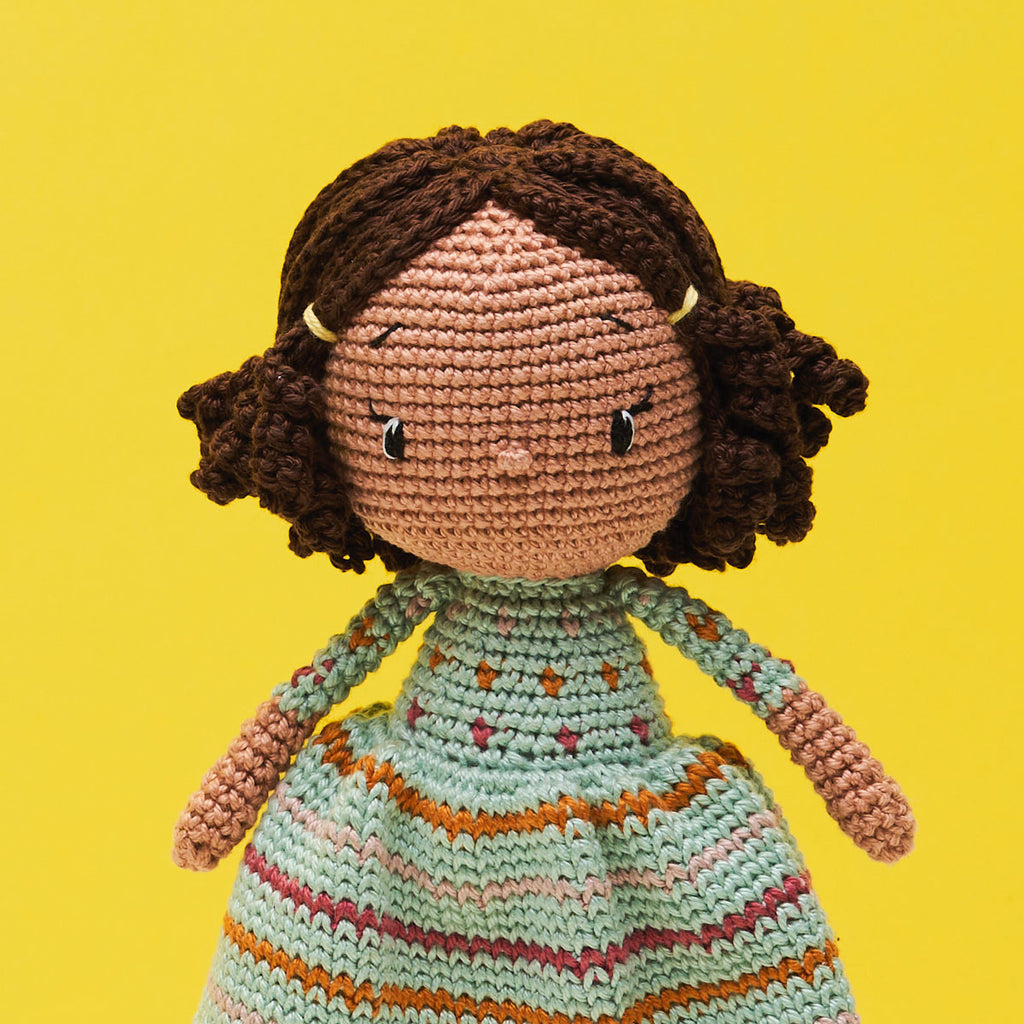 Crocheted doll with brown curly hair and a patterned dress, set against a bright yellow background. The doll exudes a cheerful, handmade charm.