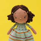 Crocheted doll with brown curly hair and a patterned dress, set against a bright yellow background. The doll exudes a cheerful, handmade charm.