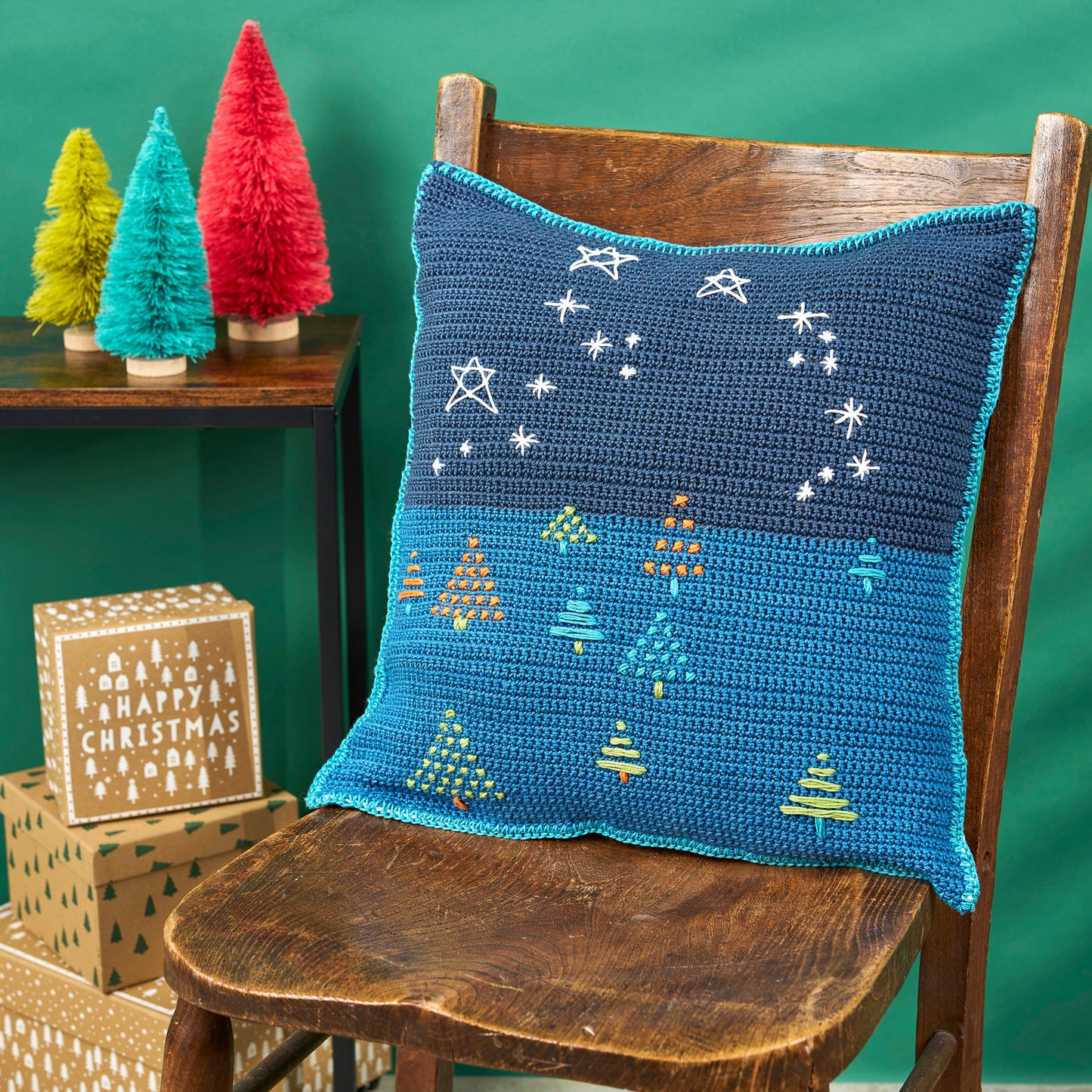 Crocheted pillow with Christmas tree design on a wooden chair against a green background.