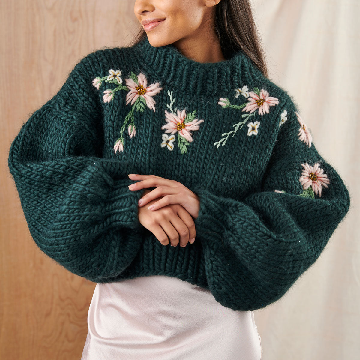 A person wearing an oversized green knitted jumper with embroidered flower details on the front.