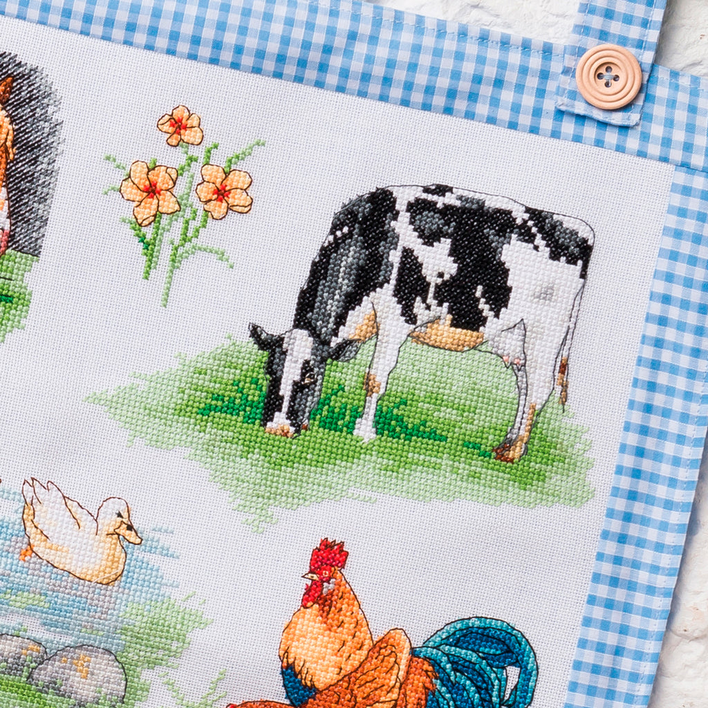 Cross-stitched panel with a cow, rooster, and duck on a blue checkered fabric background.