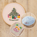 Embroidery hoop with Christmas-themed embroidery, small blue beret, and fabric patch on a wooden surface.