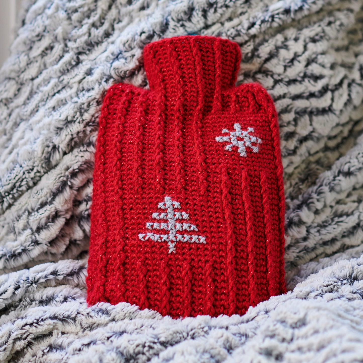 Red crocheted hot water bottle cover with a white snowflake design on a textured fabric background