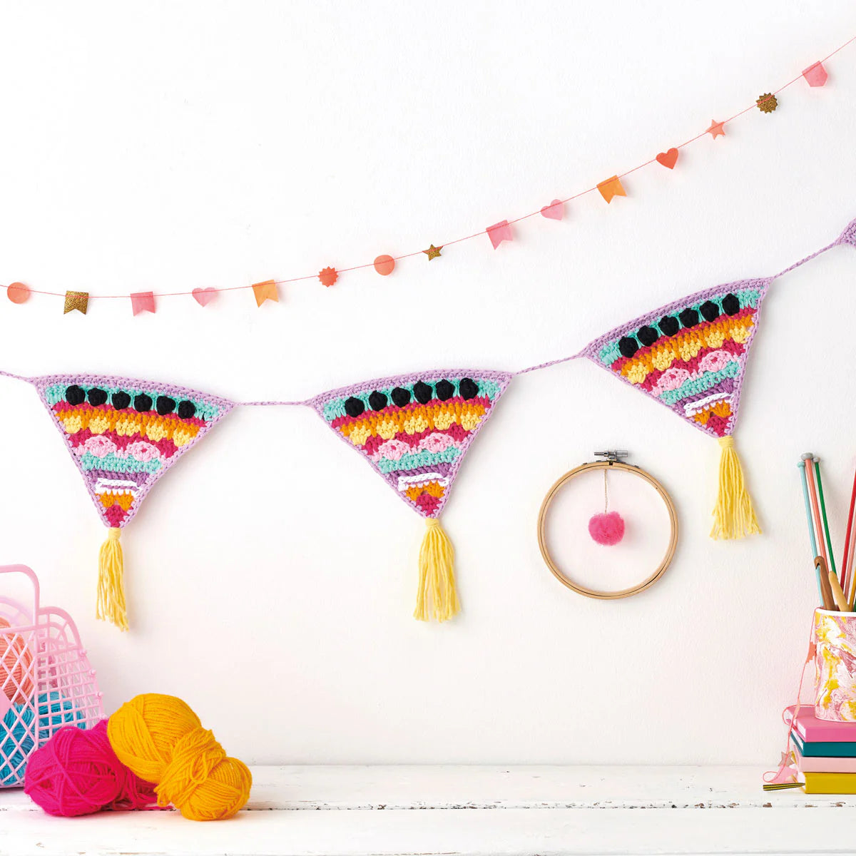 Colorful crocheted bunting with tassels on a white wall, with yarn and a hoop decoration in the foreground.
