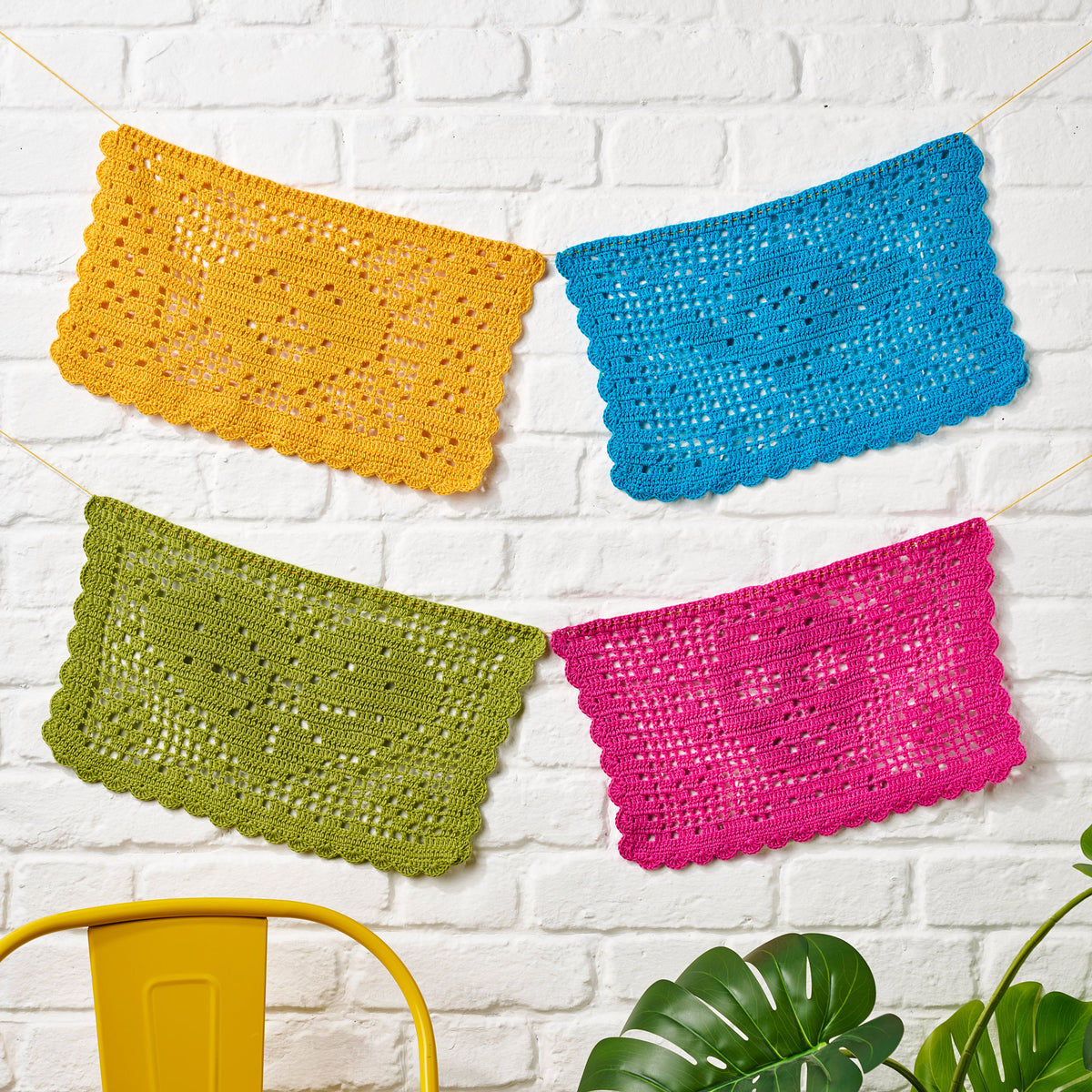Four colorful crocheted squares hanging against a white brick wall with a yellow bucket and green plant.