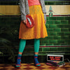 A person in a colorful, eclectic outfit stands against a vintage green tiled wall. They wear a pink top, yellow skirt, teal leggings, and striped socks. A glittery red purse with a toothy grin adds whimsy. A "Do Not Enter" sign on books enhances the playful, quirky vibe.