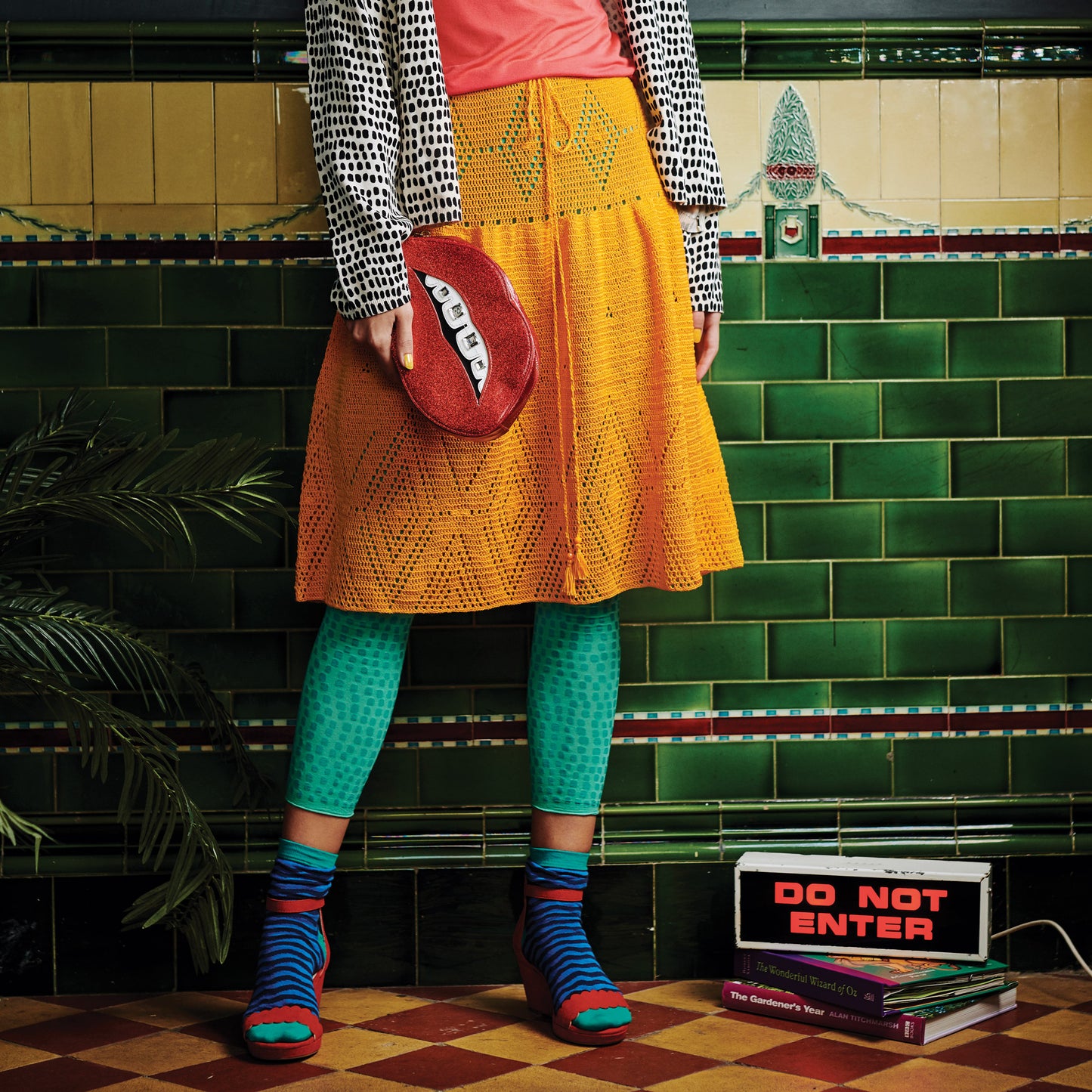 A person in a colorful, eclectic outfit stands against a vintage green tiled wall. They wear a pink top, yellow skirt, teal leggings, and striped socks. A glittery red purse with a toothy grin adds whimsy. A "Do Not Enter" sign on books enhances the playful, quirky vibe.