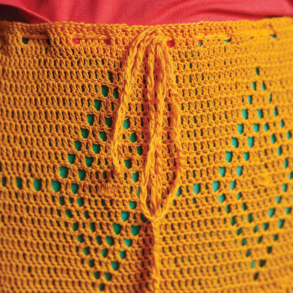 Close-up of a vibrant orange crocheted garment with a drawstring, featuring star patterns against a green background. Worn over a red fabric.