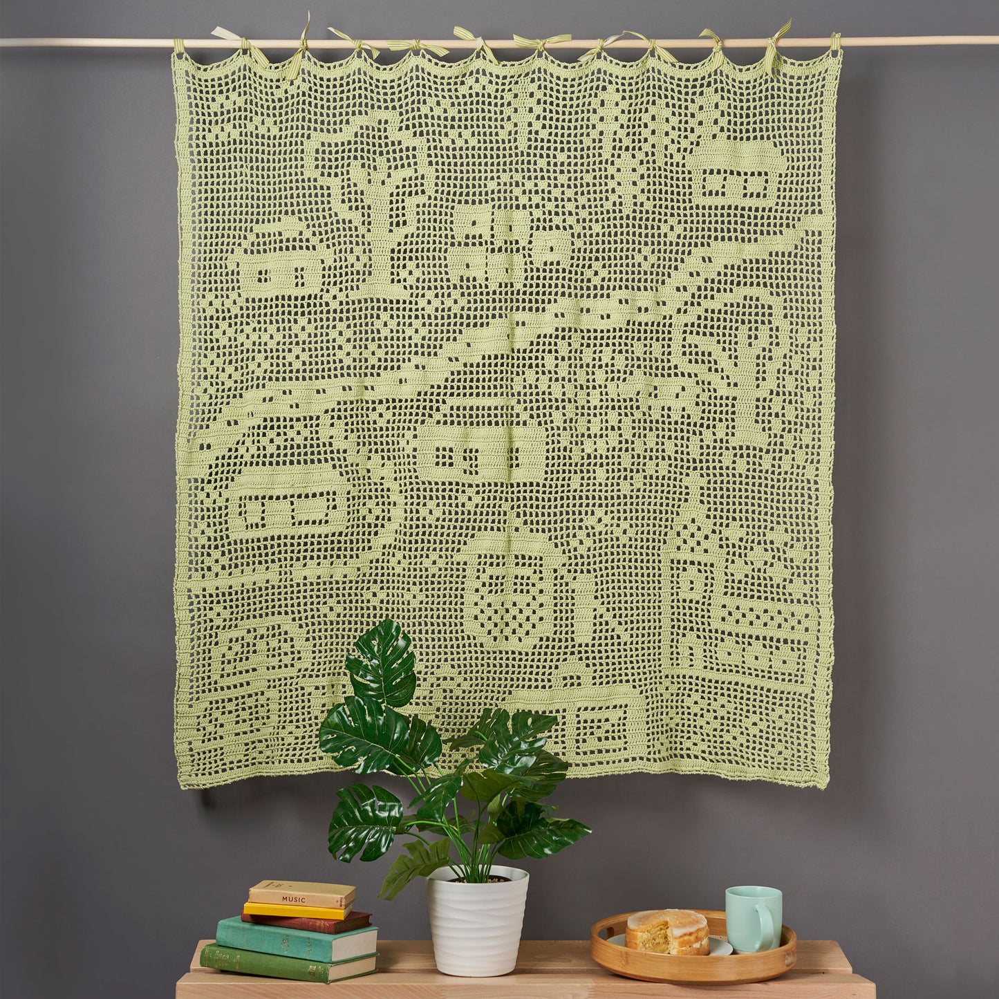 Decorative green crochet blanket with geometric pattern on a gray wall, below a wooden rod.