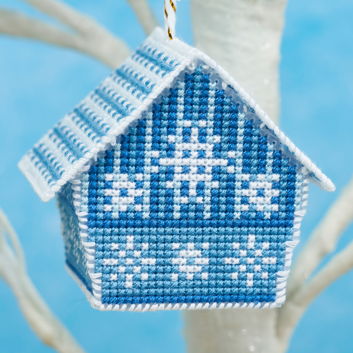 Cross-stitched blue and white house ornament on a branch with a blue background