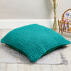 A green, knitted floor cushion placed on a white rug with a woven chair in the background.