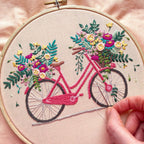Embroidery hoop with a bicycle and floral design on beige fabric.