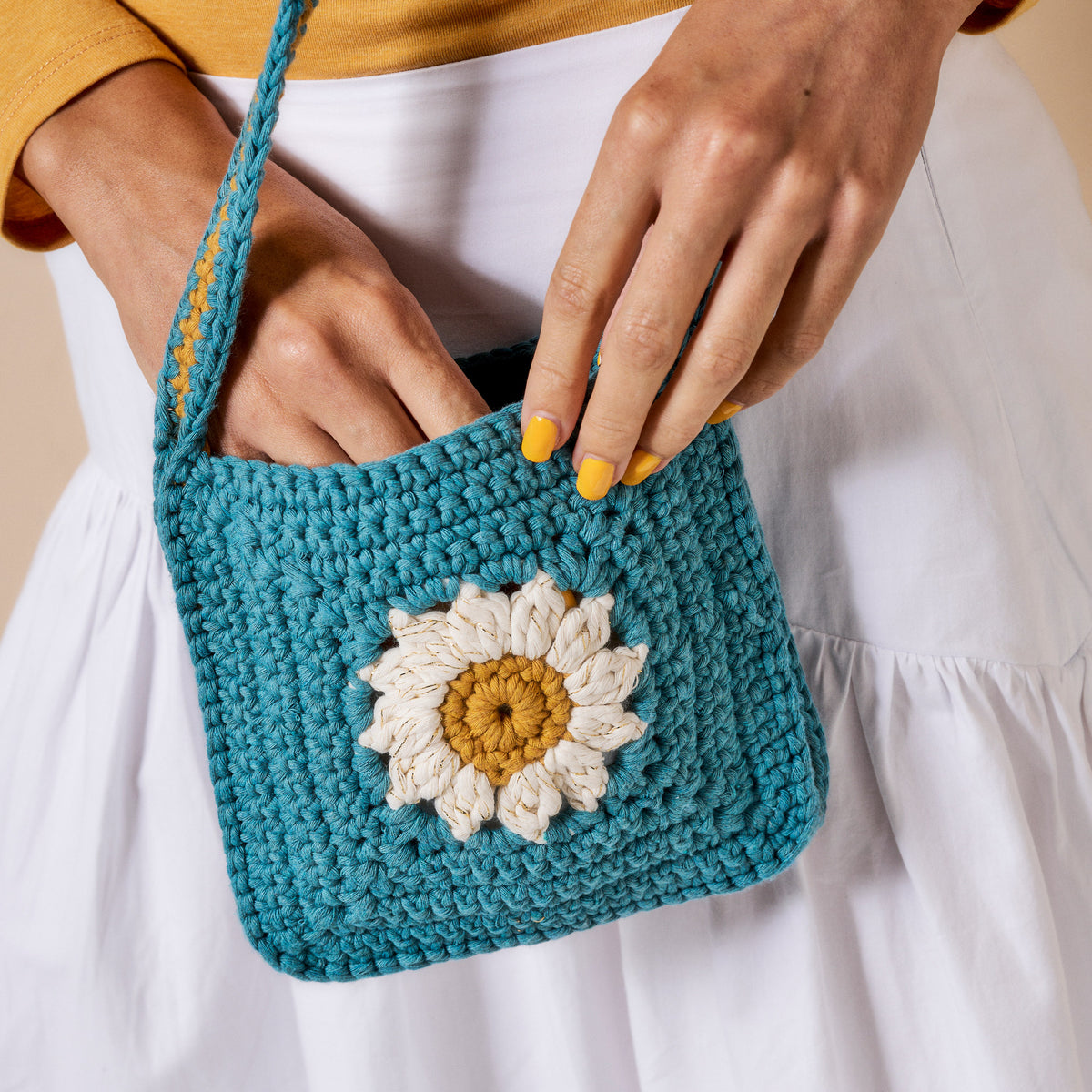 Blue crocheted purse with a floral design held by a person wearing a yellow top and white pants.