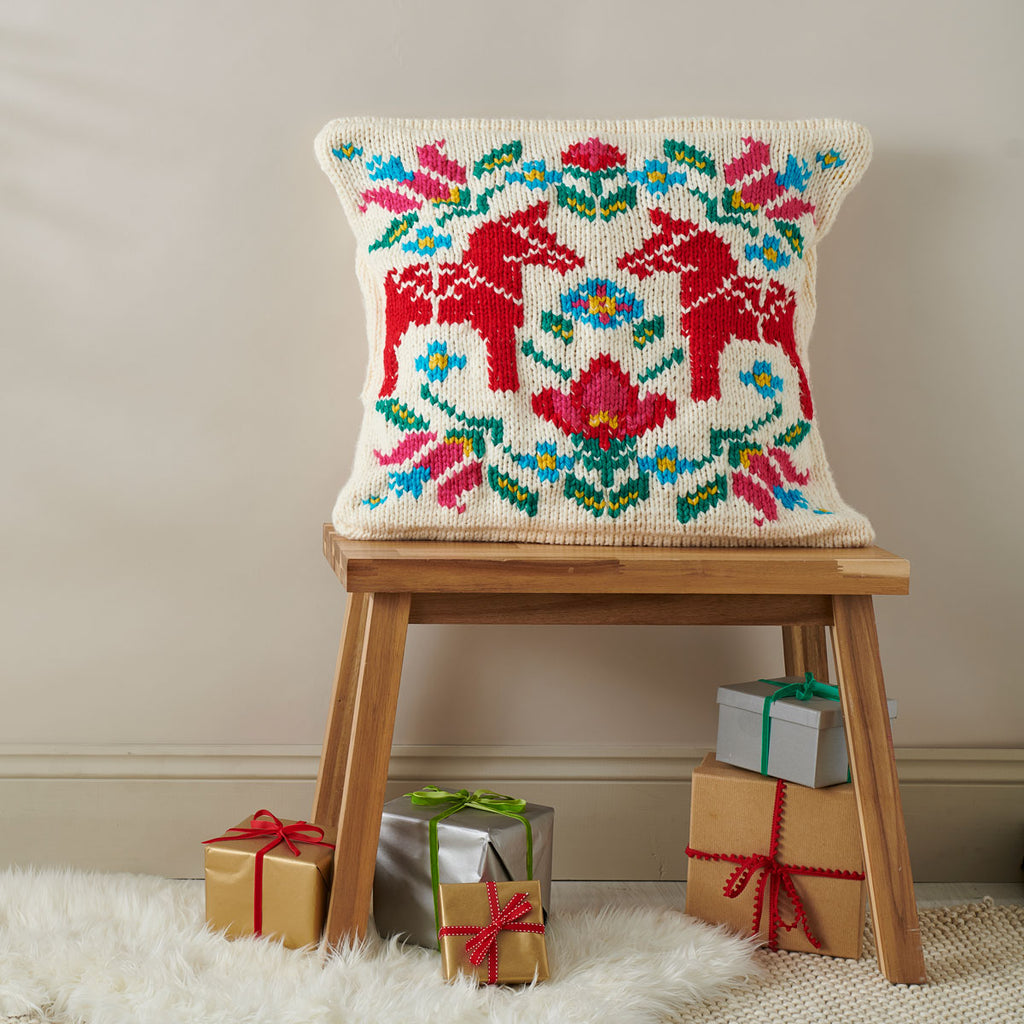 A knitted cushion with a colorful folk art design featuring geometric patterns and the Swedish Dala horse, displayed on a wooden stool beside wrapped gifts.