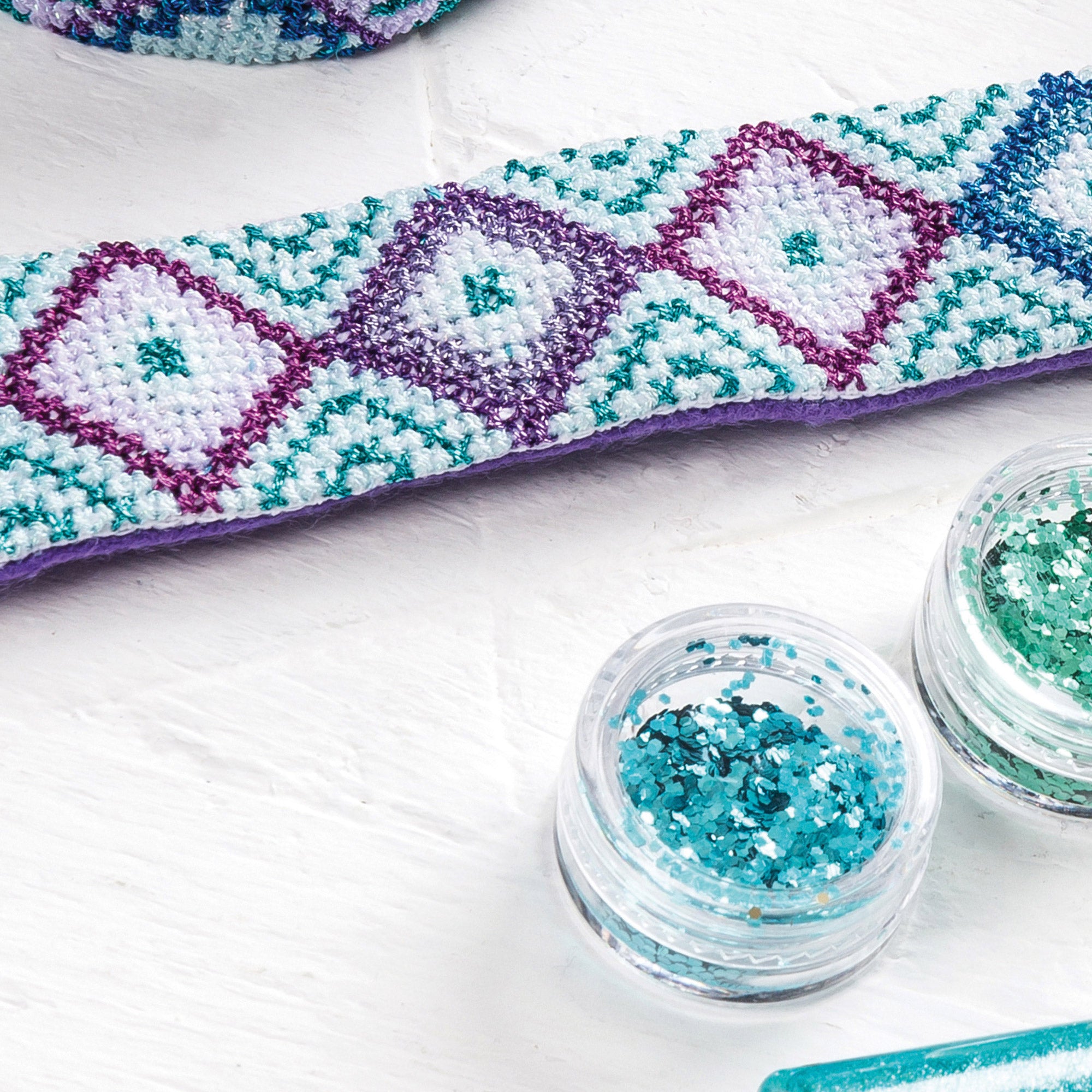 Cross-stitched heart design with two containers of glitter on a white surface