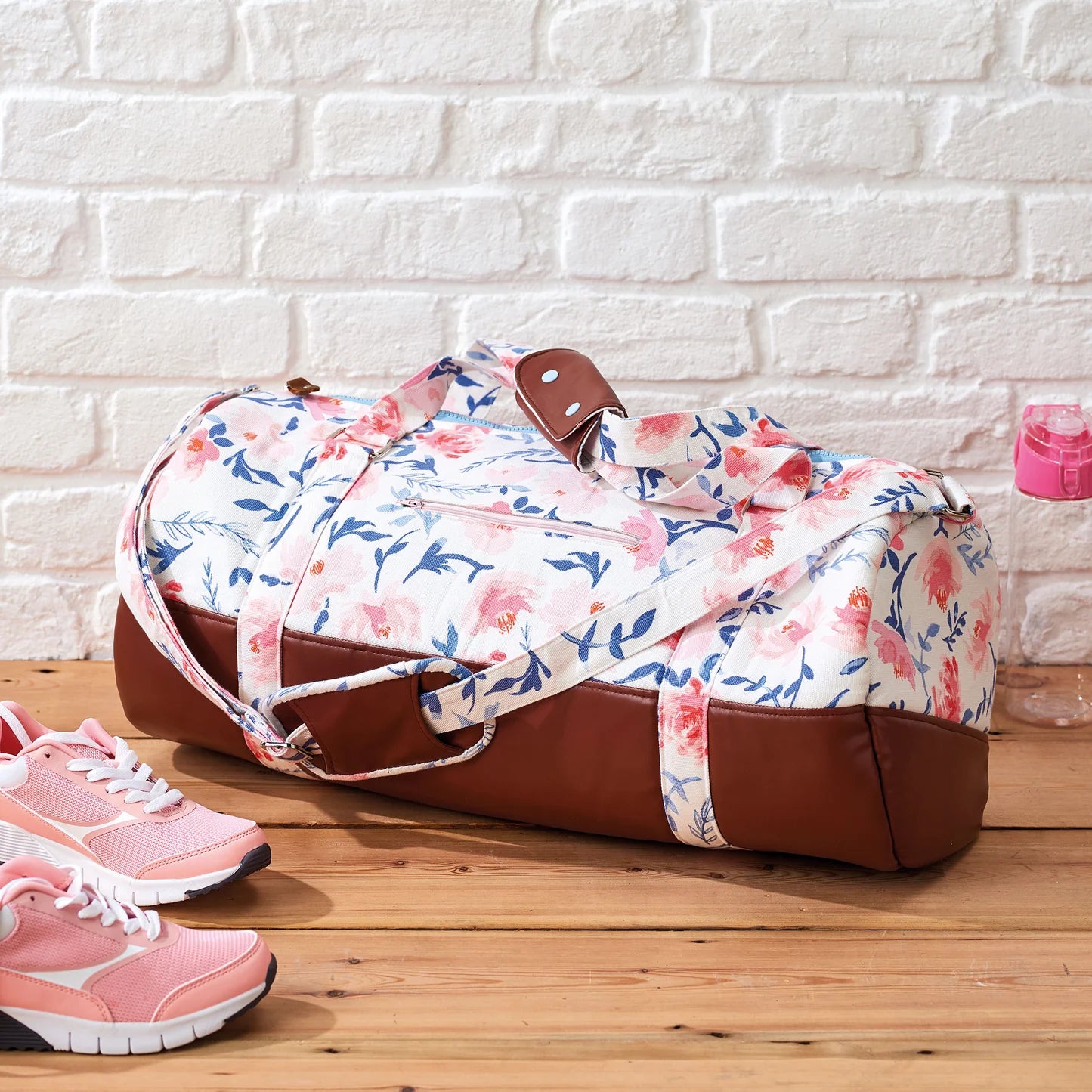 Floral-patterned duffel bag with brown accents on a wooden floor against a white brick wall.