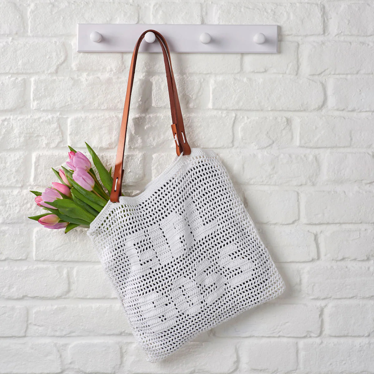 White crochet bag with brown handles holding pink tulips, hanging on a white brick wall.