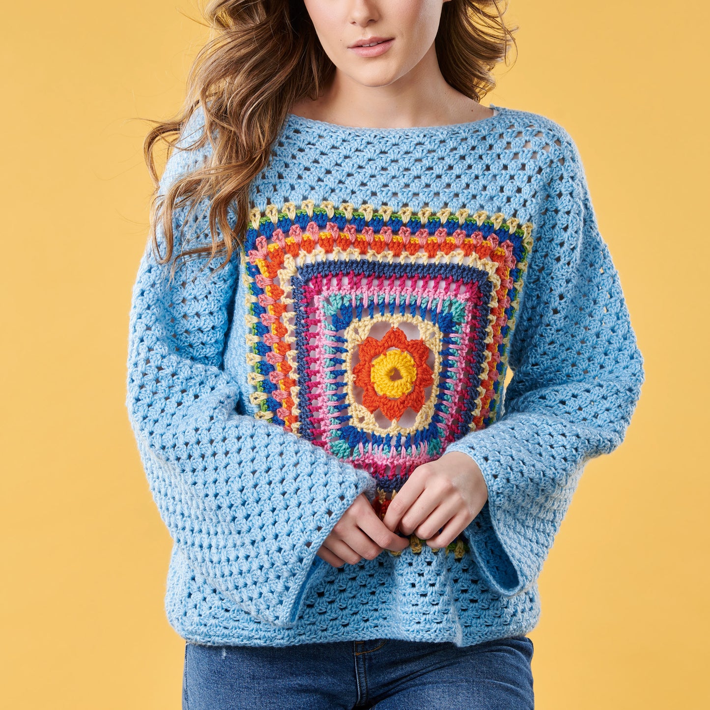 Person wearing a blue crochet sweater with a colorful square pattern on a yellow background
