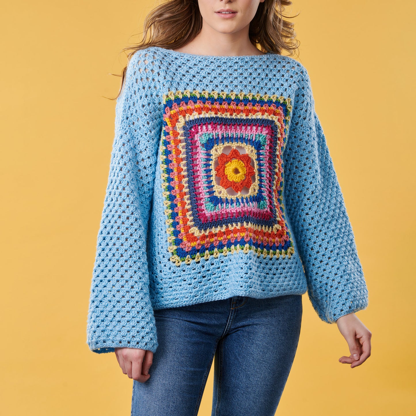 Person wearing a blue crochet sweater with a colorful square pattern on a yellow background