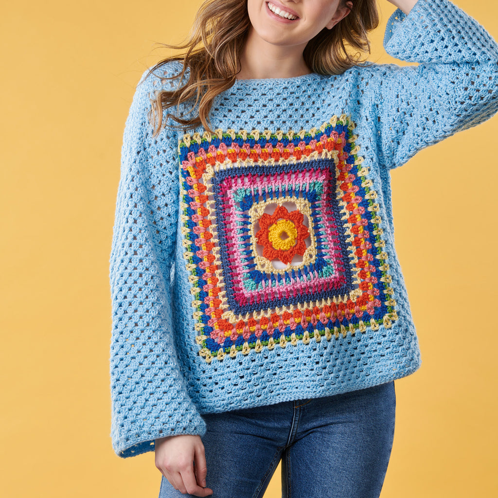 Person wearing a blue crochet sweater with a colorful square pattern on a yellow background