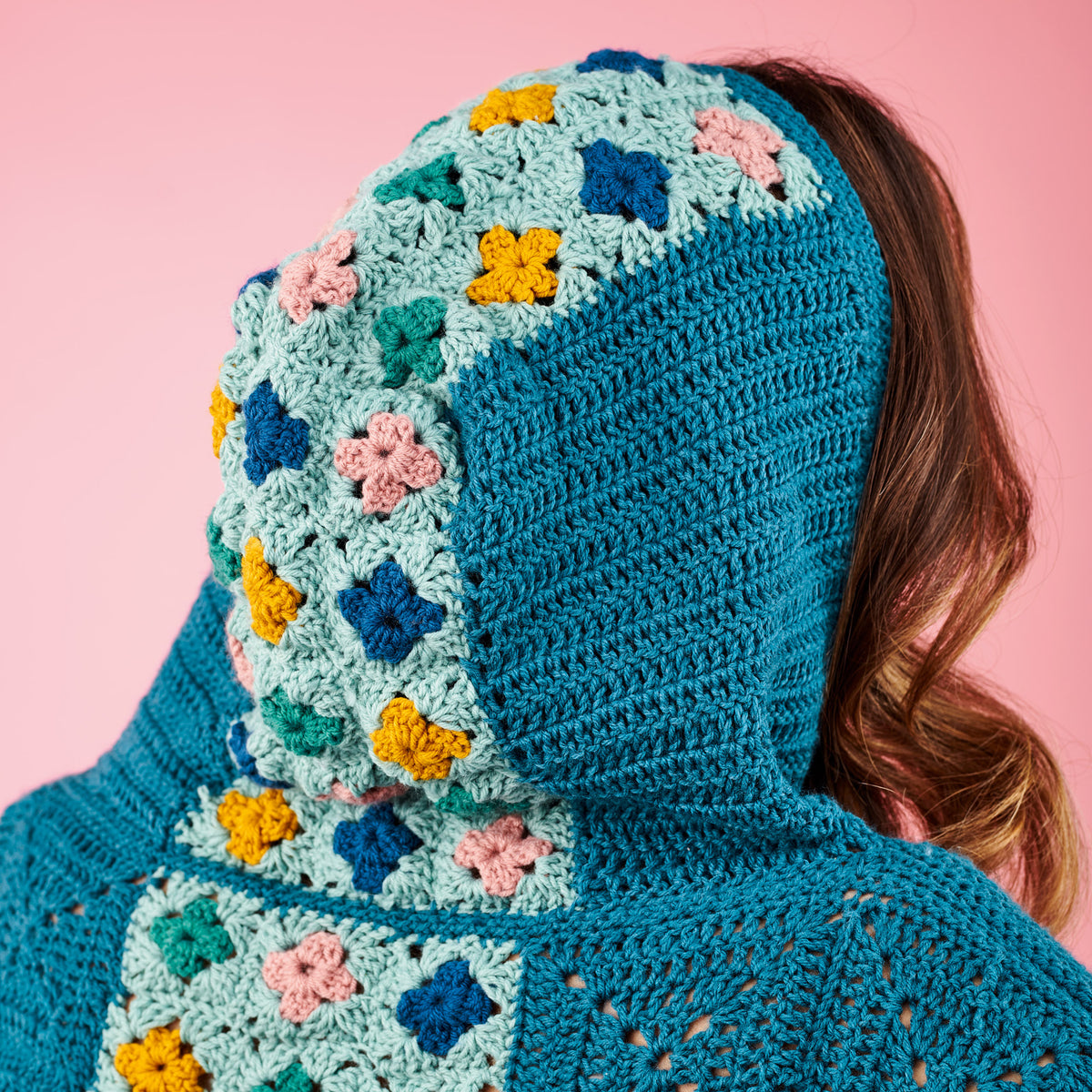 Person wearing a blue crocheted hoodie with floral patterns on a pink background