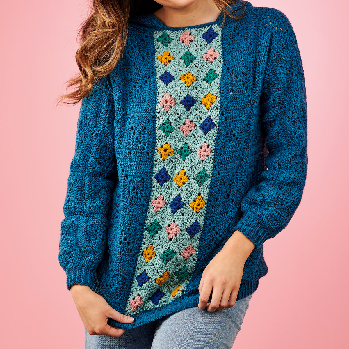 Person wearing a blue crochet cardigan with a colorful pattern on a pink background
