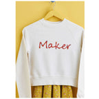 A white sweatshirt with the word "Maker" in colorful letters hangs on a wooden hanger. Below, a yellow floral skirt peeks out, against a yellow background.