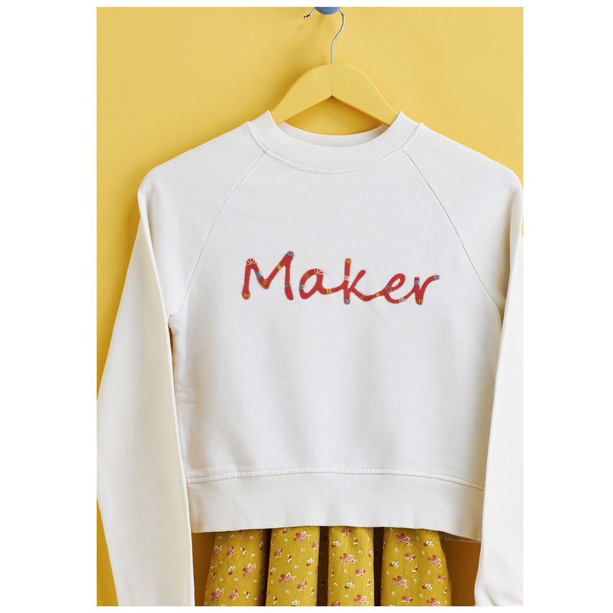 A white sweatshirt with the word "Maker" in colorful letters hangs on a wooden hanger. Below, a yellow floral skirt peeks out, against a yellow background.