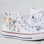 White high-top sneakers featuring colorful floral embroidery and red accents on the soles. The Converse logo is visible, conveying a stylish, creative vibe.