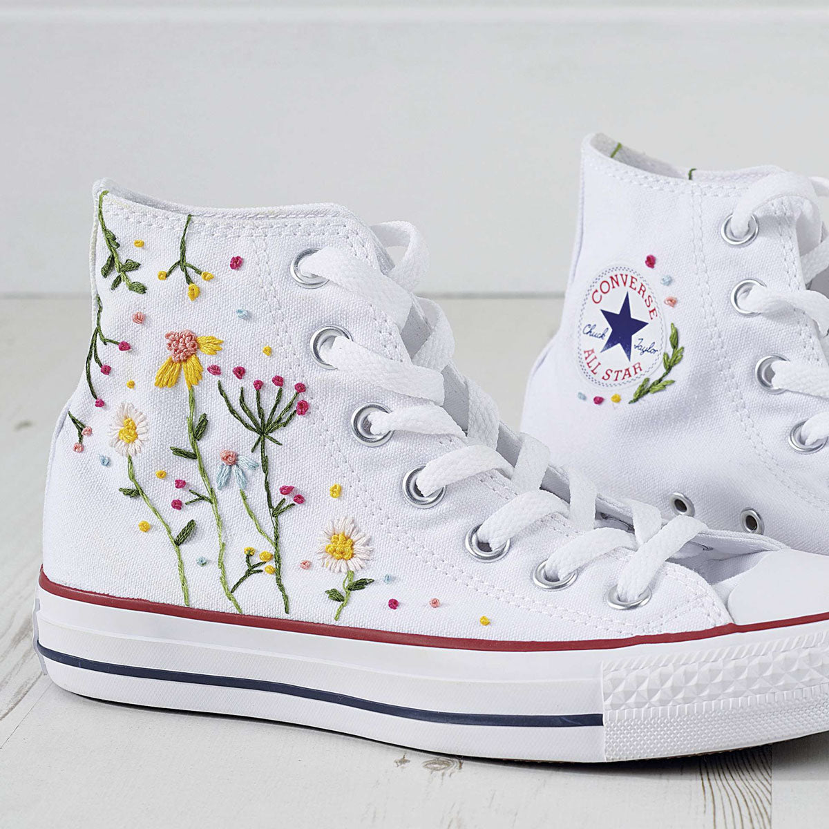 White high-top sneakers featuring colorful floral embroidery and red accents on the soles. The Converse logo is visible, conveying a stylish, creative vibe.