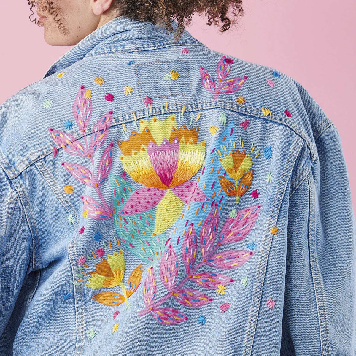 A person with curly hair wears a blue denim jacket embroidered with colorful, vibrant flowers and leaves. The background is a soft pink.