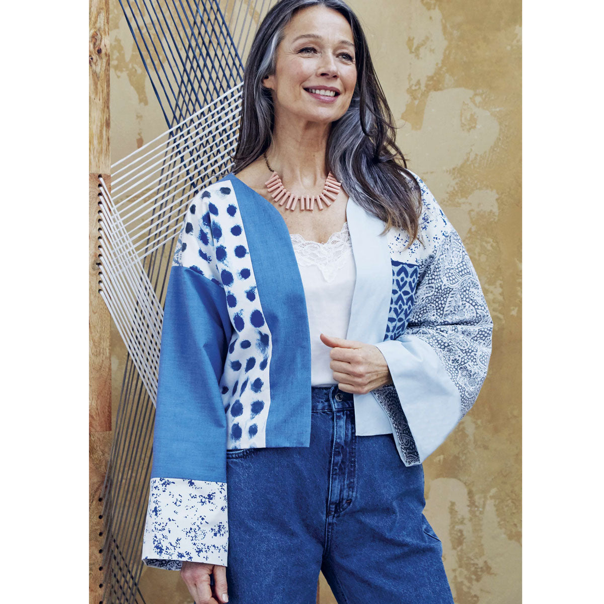 Smiling woman in a patchwork kimono with blue patterns, white lace top, and jeans. She stands in front of a textured wall with woven strings, exuding charm and warmth.