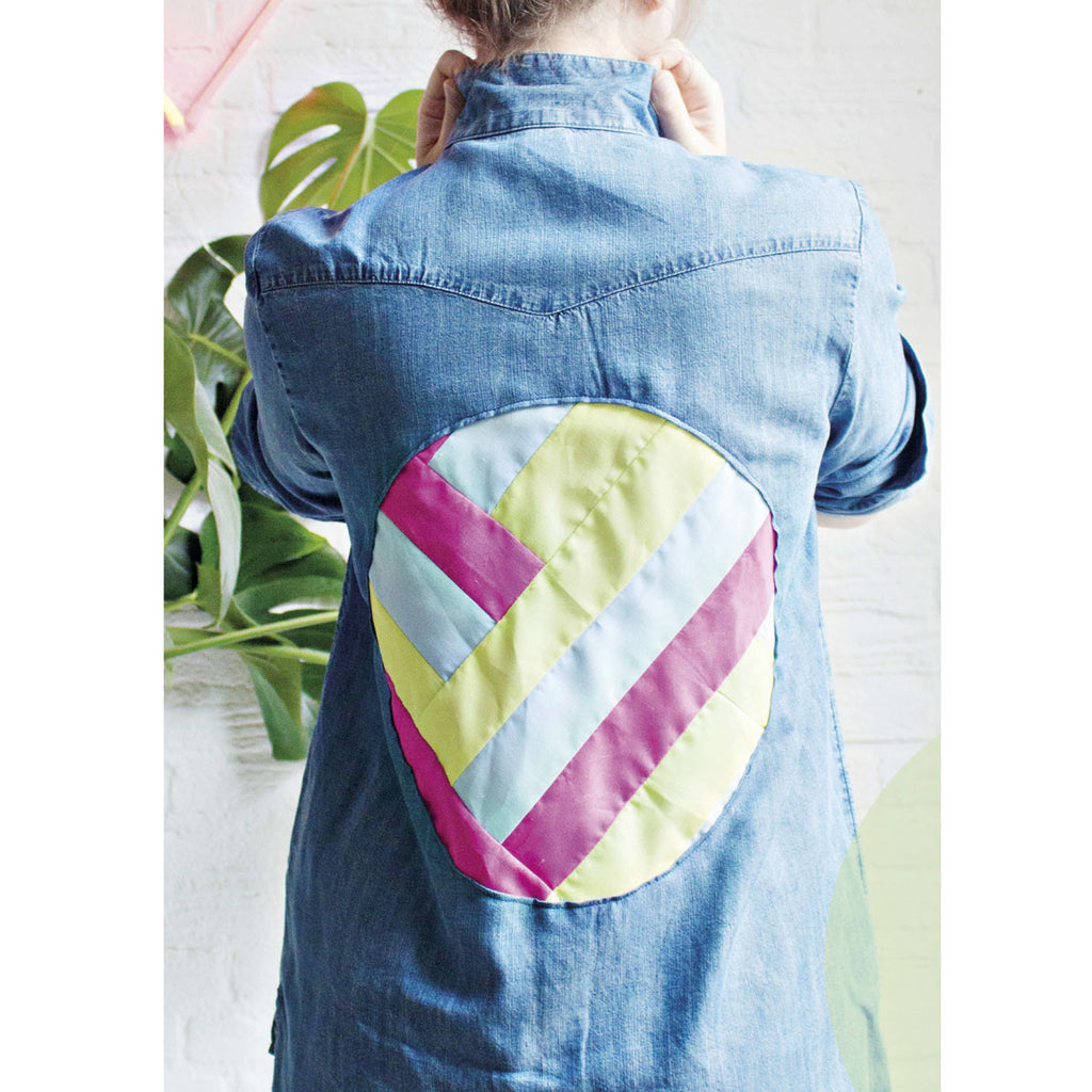 A person wearing a denim jacket with a colorful, quilted geometric patch on the back, featuring pink, yellow, and blue stripes, stands in front of a plant.