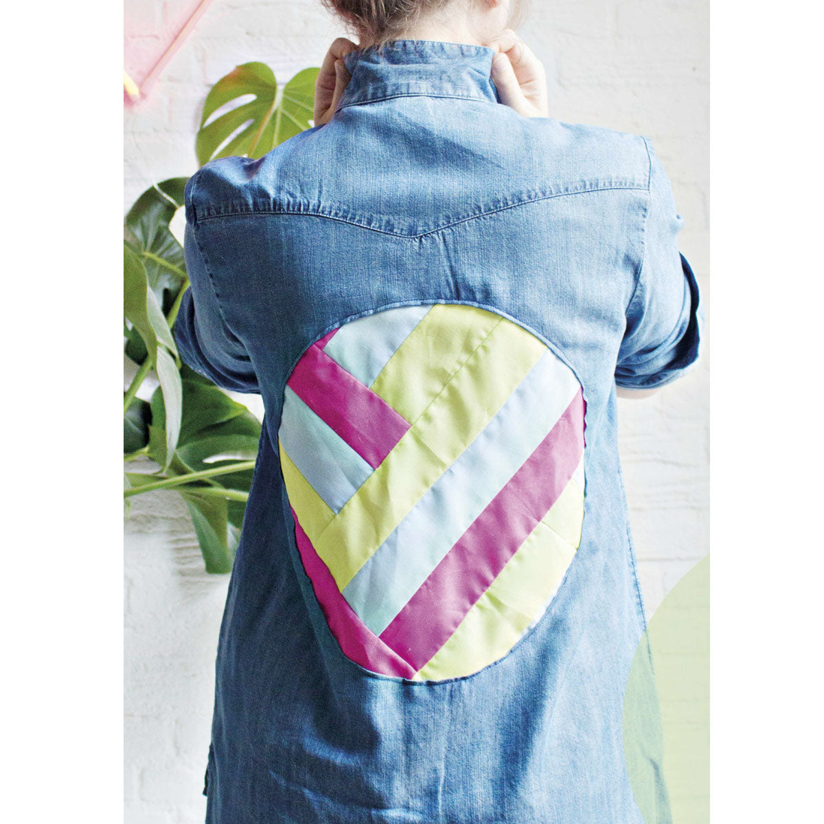 A person wearing a denim jacket with a colorful, quilted geometric patch on the back, featuring pink, yellow, and blue stripes, stands in front of a plant.