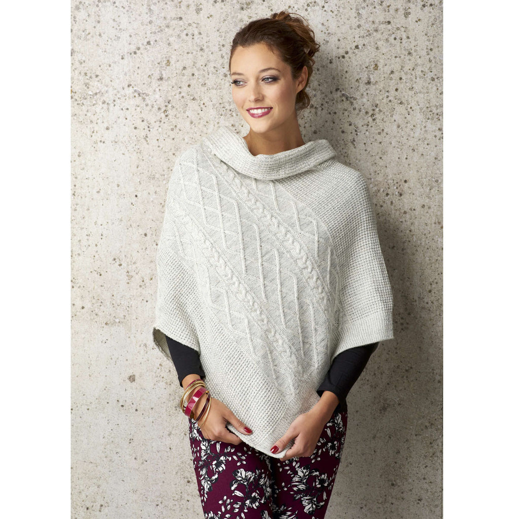 Woman in a cozy cream cable-knit poncho, smiling and gazing to the side against a textured stone wall. She wears floral pants and colorful bangles.