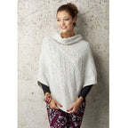 Woman in a cozy cream cable-knit poncho, smiling and gazing to the side against a textured stone wall. She wears floral pants and colorful bangles.