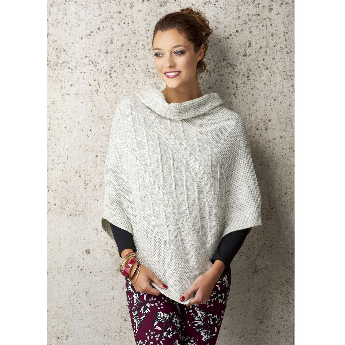 Woman in a cozy cream cable-knit poncho, smiling and gazing to the side against a textured stone wall. She wears floral pants and colorful bangles.