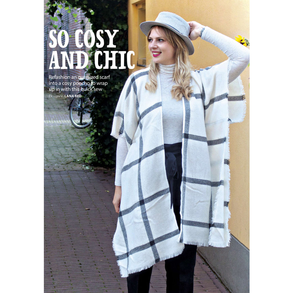A woman stands on a cobblestone street wearing a cozy, chic poncho with gray and white checks. She accessorizes with a gray hat, conveying a stylish, autumn vibe.