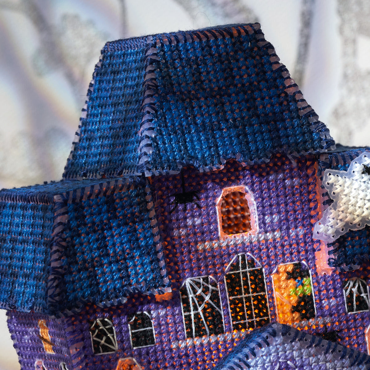Cross-stitched miniature house with a blue roof and purple body on a textured white background