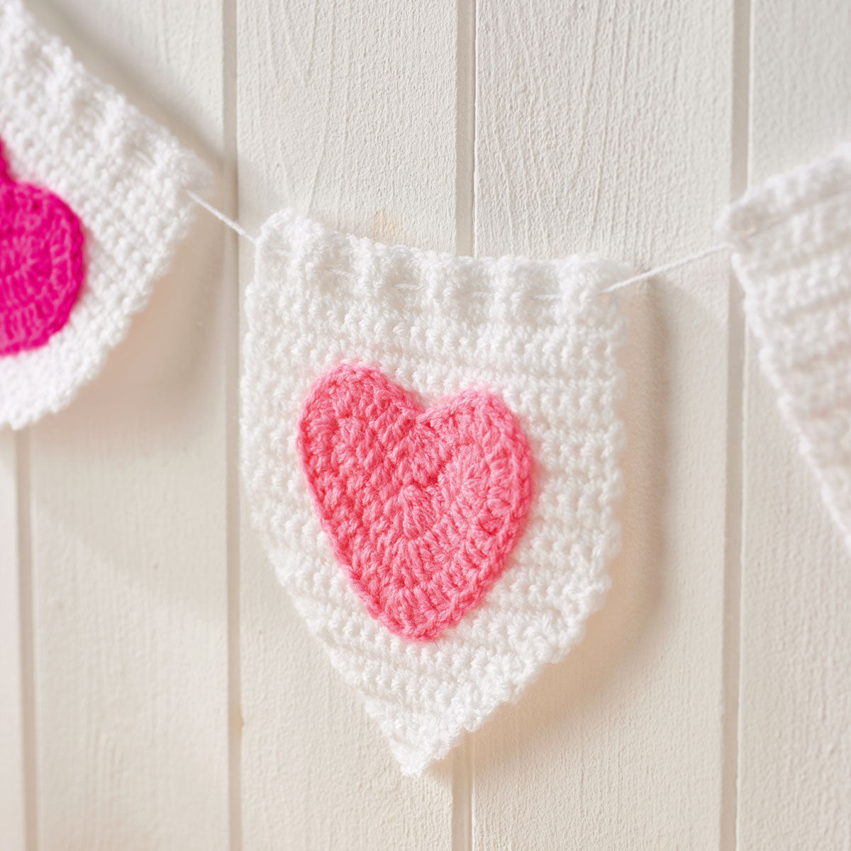 Crocheted white buntings with pink hearts hang against a wooden wall. The texture is cozy, creating a warm and cheerful atmosphere.