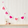 A string of crochet bunting with pink heart shapes on a white pennant, displayed on a wooden shelf with a spool of thread and scissors.