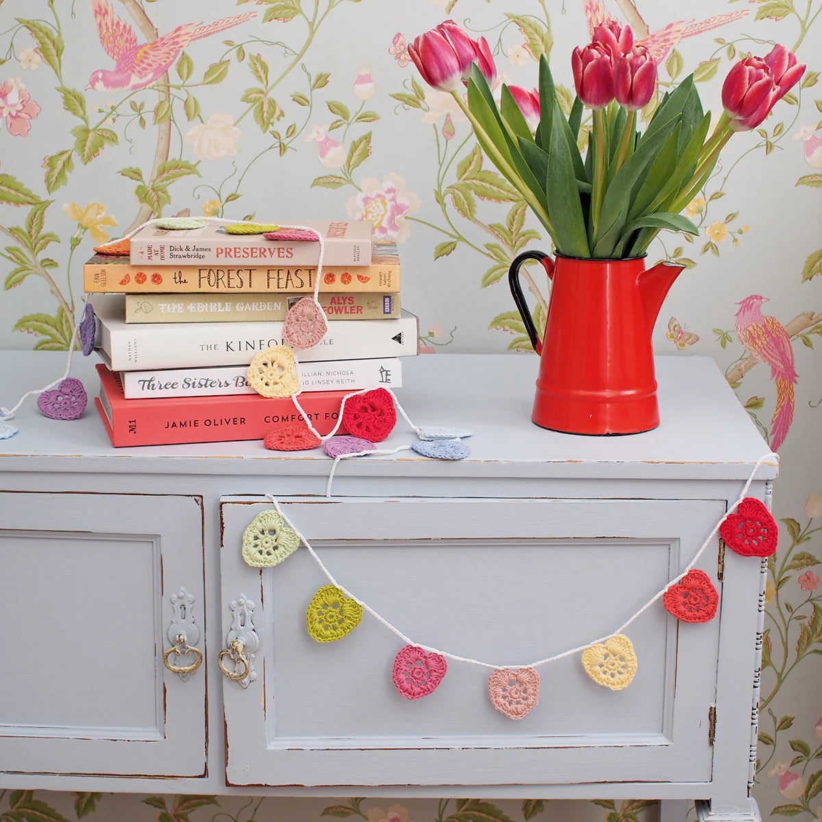 White cabinet with books and a red vase of tulips against floral wallpaper