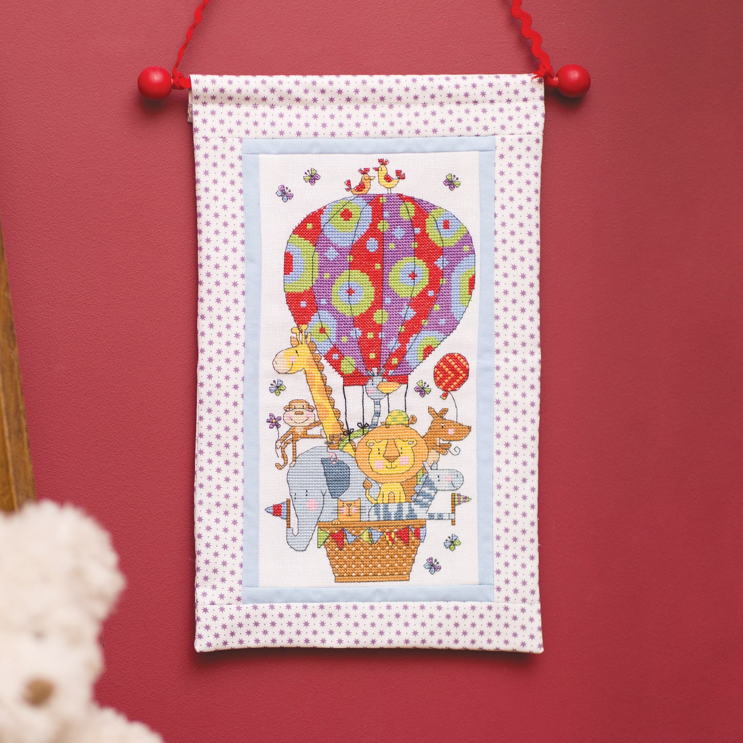 Cross-stitched panel with colorful hot air balloon and animals on a polka dot background, hanging against a red wall.